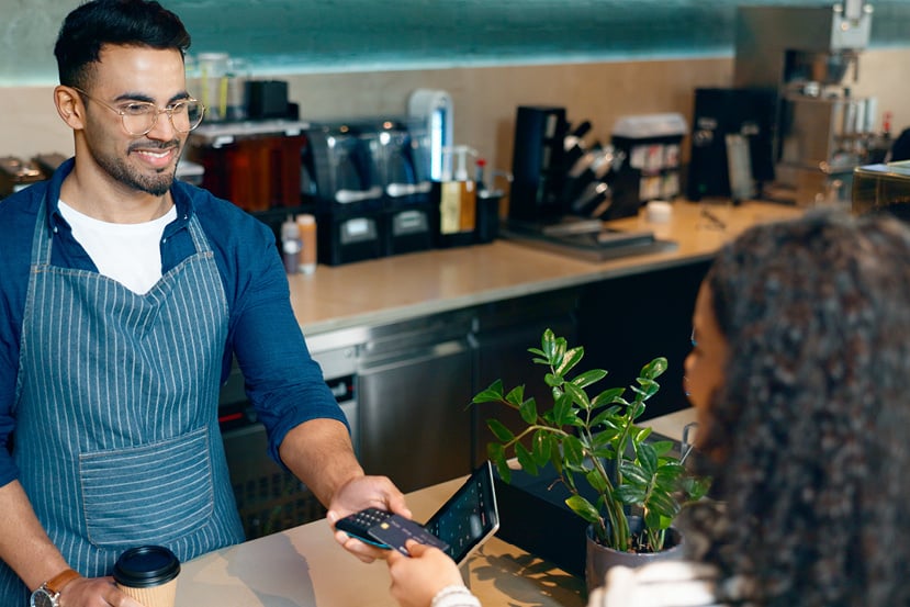 A young man cashier accepting payment on a wireless card reader at a coffee shop.