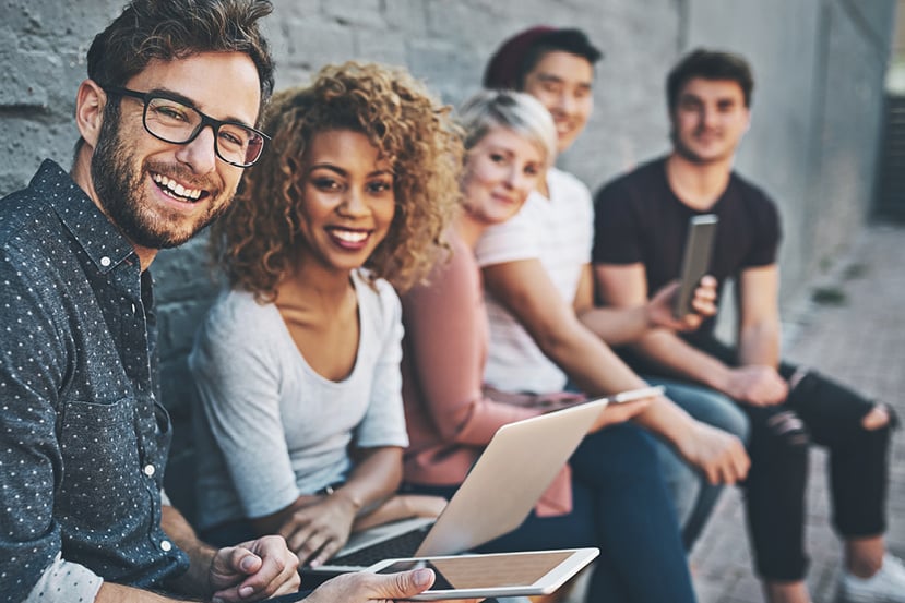 Group of young adults sitting together outdoors, smiling and using laptops and tablets.